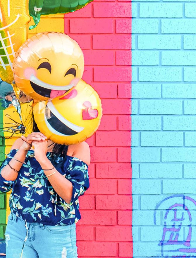 Frau mit mehreren Emoji-Luftballons steht vor einer buntgestreiften Wand.