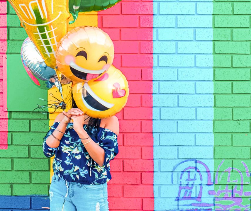 Frau mit mehreren Emoji-Luftballons steht vor einer buntgestreiften Wand.