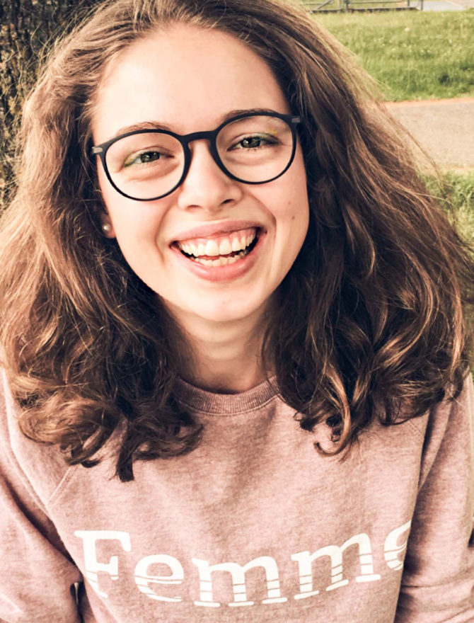 Junge Frau mit braunen Haaren und großer Brille mit Femme-Pullover.