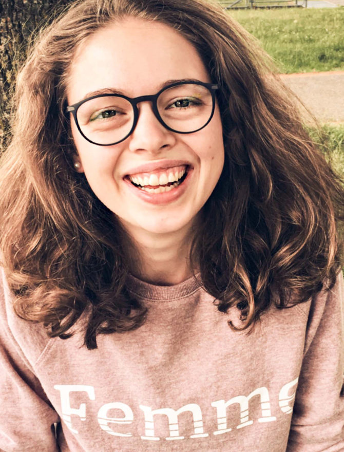 Junge Frau mit braunen Haaren und großer Brille mit Femme-Pullover.