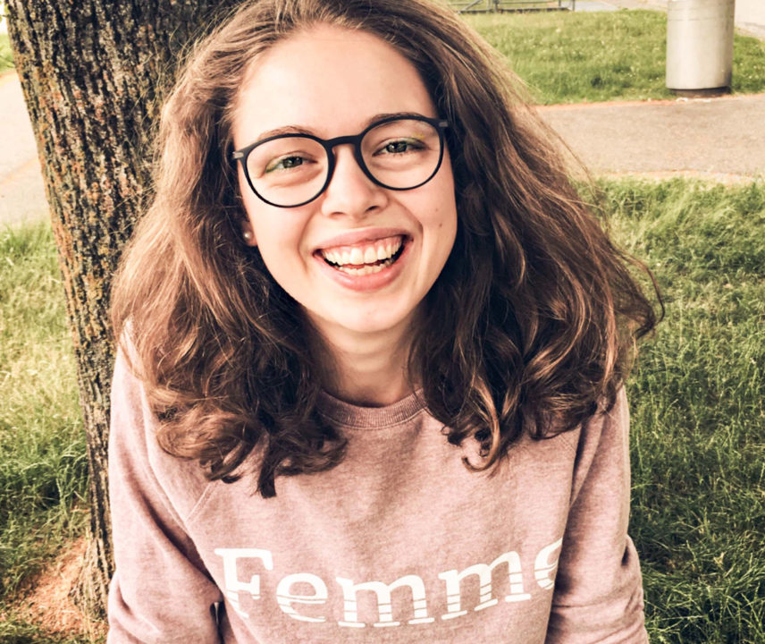 Junge Frau mit braunen Haaren und großer Brille mit Femme-Pullover.