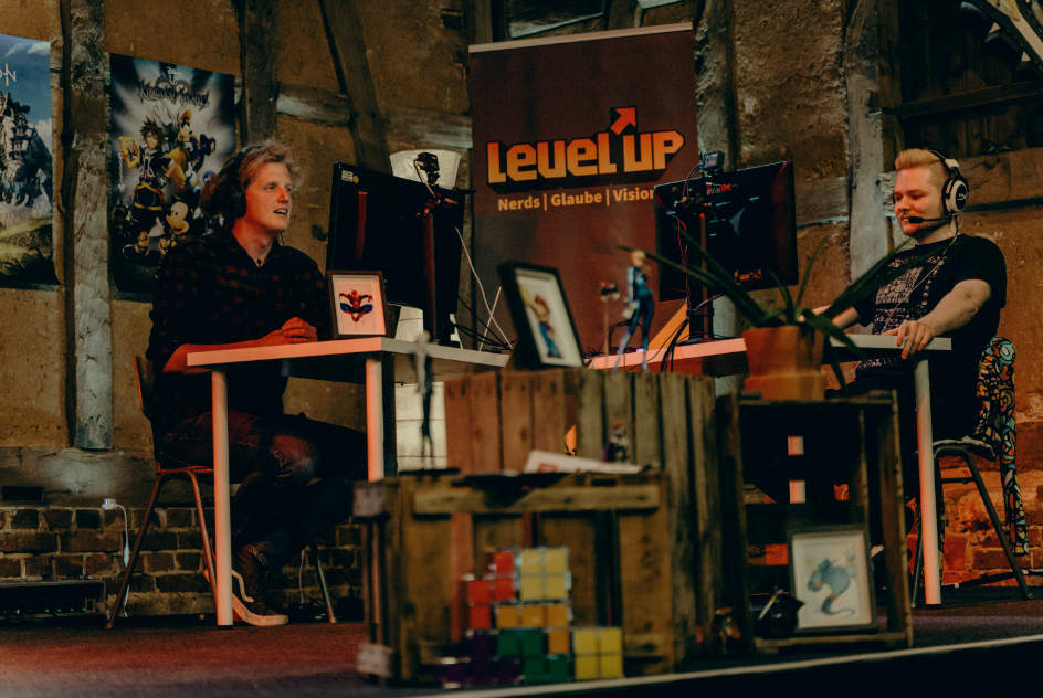 Zwei Junge Menschen an ihren Rechnern. Im Hintergrund Dekoration im Gamer-Stil für die Level'up-Konferenz.