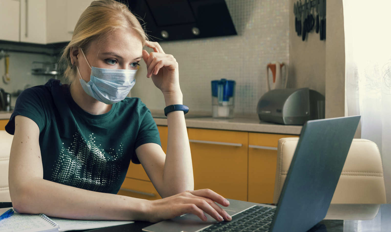 Studentin in der Küche mit medizinischer Maske am Laptop
