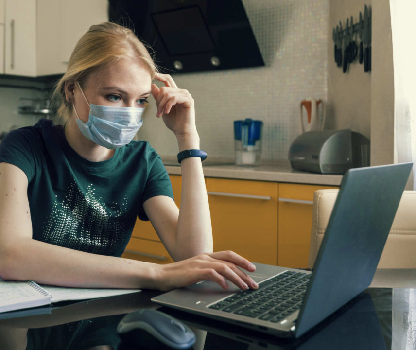 Studentin in der Küche mit medizinischer Maske am Laptop