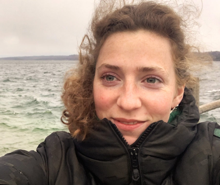 Junge Frau mit blonden lockigen Haaren und schwarzer Winterjacke vom Wind verweht am Meer