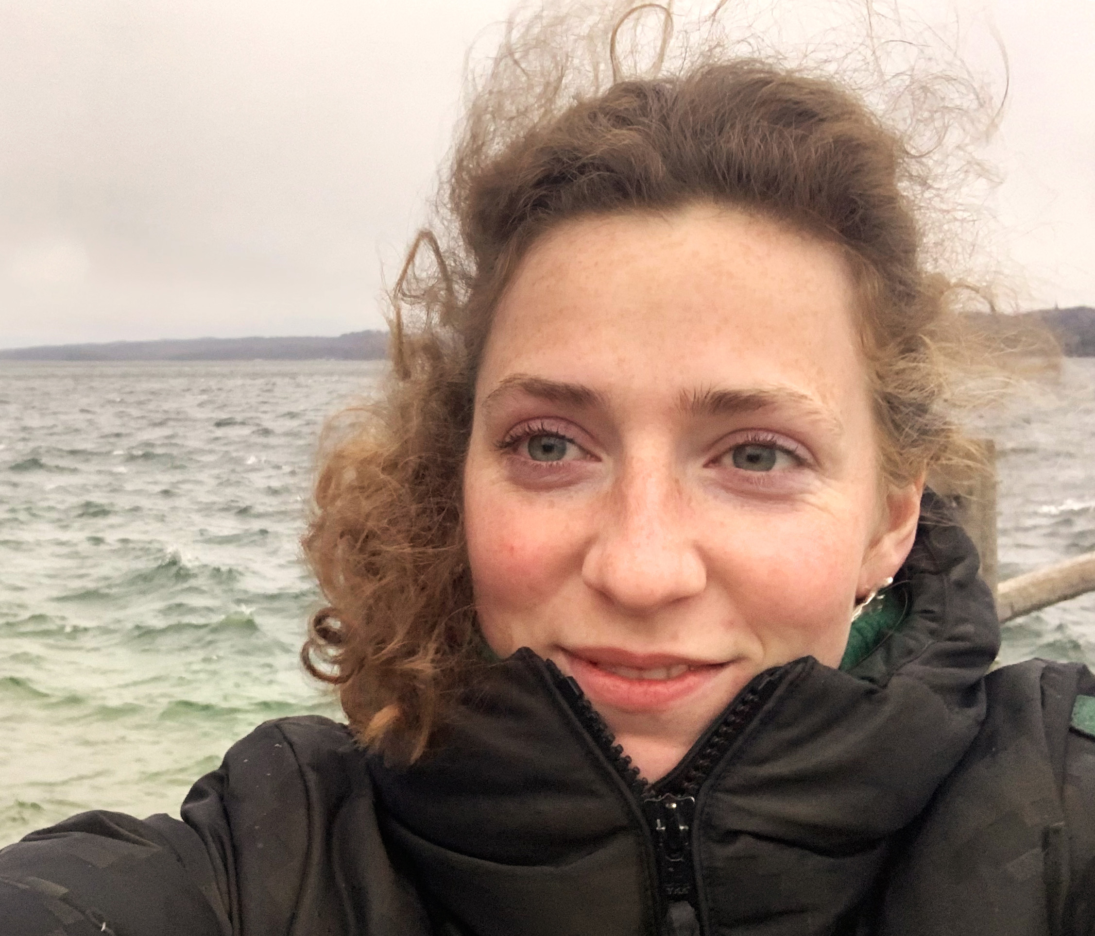 Junge Frau mit blonden lockigen Haaren und schwarzer Winterjacke vom Wind verweht am Meer