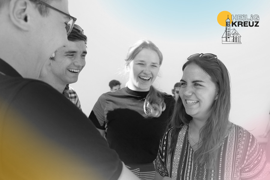 Junge Erwachsene begrüßen sich lachend in schwarz-weiß mit Logo der Heilig-Kreuz-Kirche Augsburg