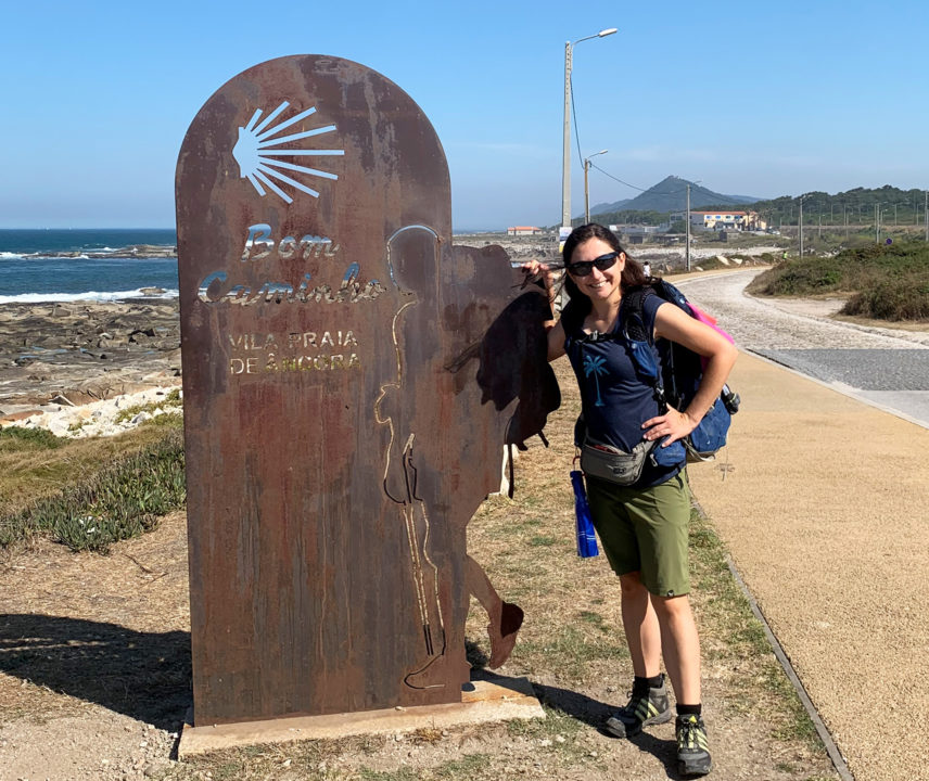 Junge dunkelhaarige Frau mit Rucksack an einer Wegmarke des Jakobswegs am Meer