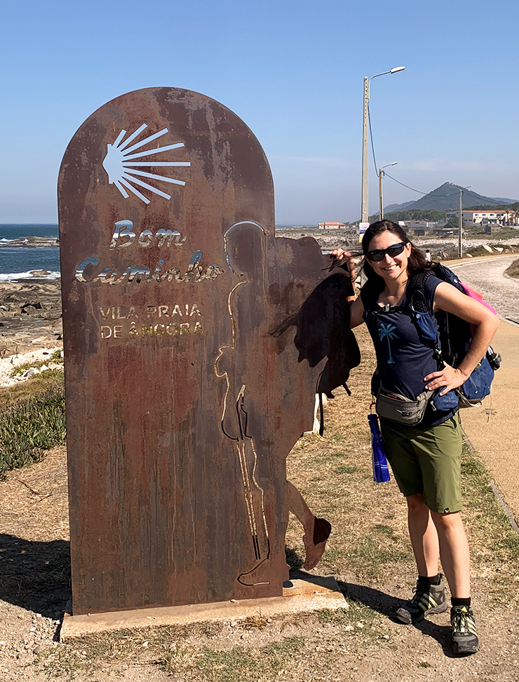 Junge dunkelhaarige Frau mit Rucksack an einer Wegmarke des Jakobswegs am Meer