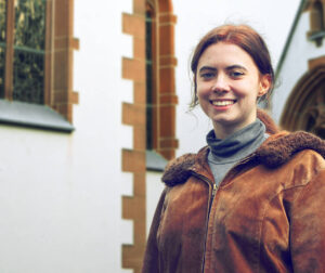 Junge Frau mit zusammengebundenen brünetten Haaren in brauner Winter-Lederjacke vor einer Kirche