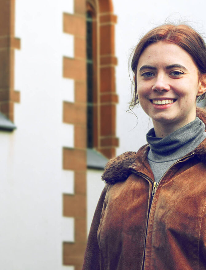 Junge Frau mit zusammengebundenen brünetten Haaren in brauner Winter-Lederjacke vor einer Kirche