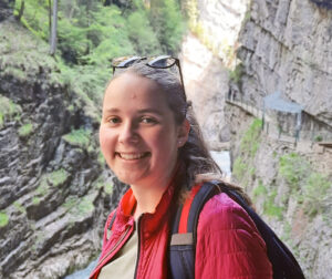 Junge Frau mit blonden Haaren und Sonnenbrille in roter Jacke und mit Rucksack in einer Schlucht