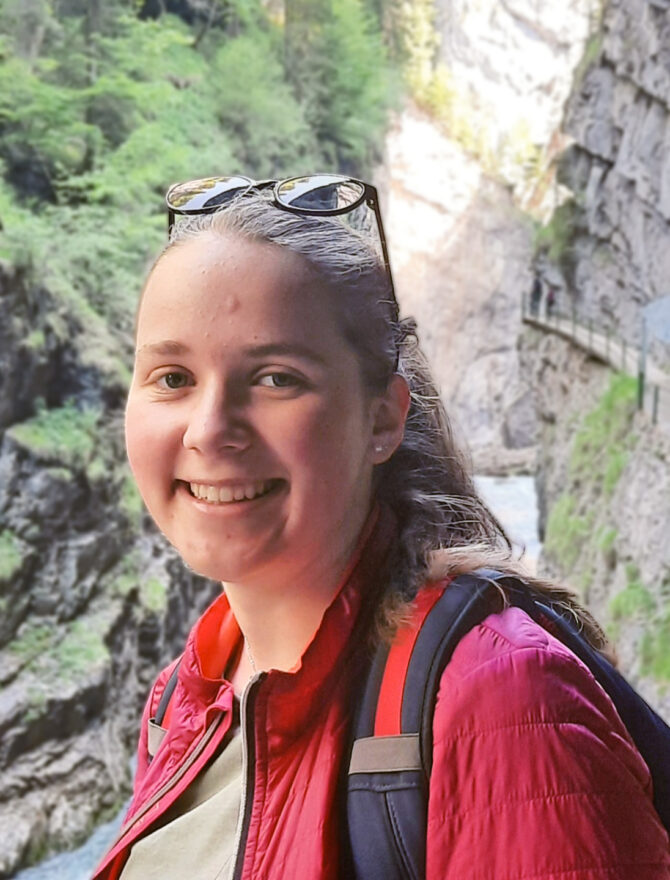 Junge Frau mit blonden Haaren und Sonnenbrille in roter Jacke und mit Rucksack in einer Schlucht