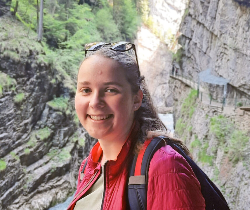 Junge Frau mit blonden Haaren und Sonnenbrille in roter Jacke und mit Rucksack in einer Schlucht
