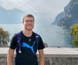 Junger europäischer Mann mit kurz blonden Haaren, Bart, schwarzer rechteckiger Brille in schwarzem T-Shirt und silberner Kreuzkette vor schöner Aussicht mit Bergen und See