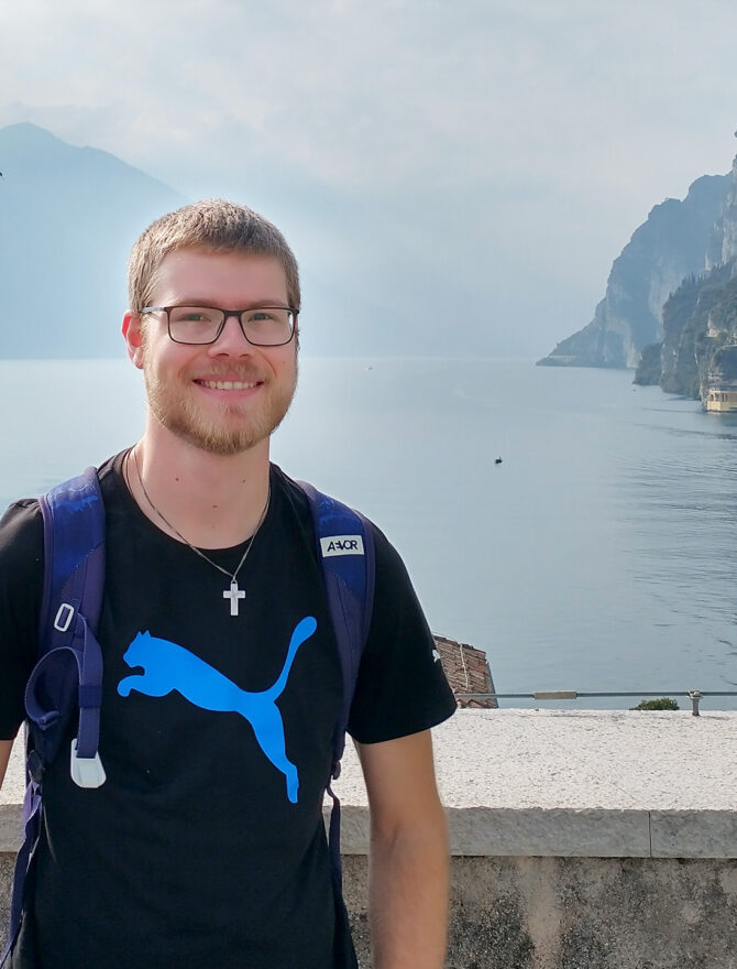 Junger europäischer Mann mit kurz blonden Haaren, Bart, schwarzer rechteckiger Brille in schwarzem T-Shirt und silberner Kreuzkette vor schöner Aussicht mit Bergen und See