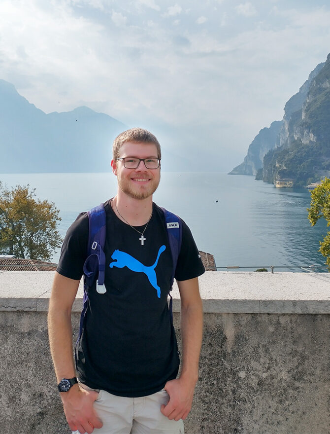 Junger europäischer Mann mit kurz blonden Haaren, Bart, schwarzer rechteckiger Brille in schwarzem T-Shirt und silberner Kreuzkette vor schöner Aussicht mit Bergen und See