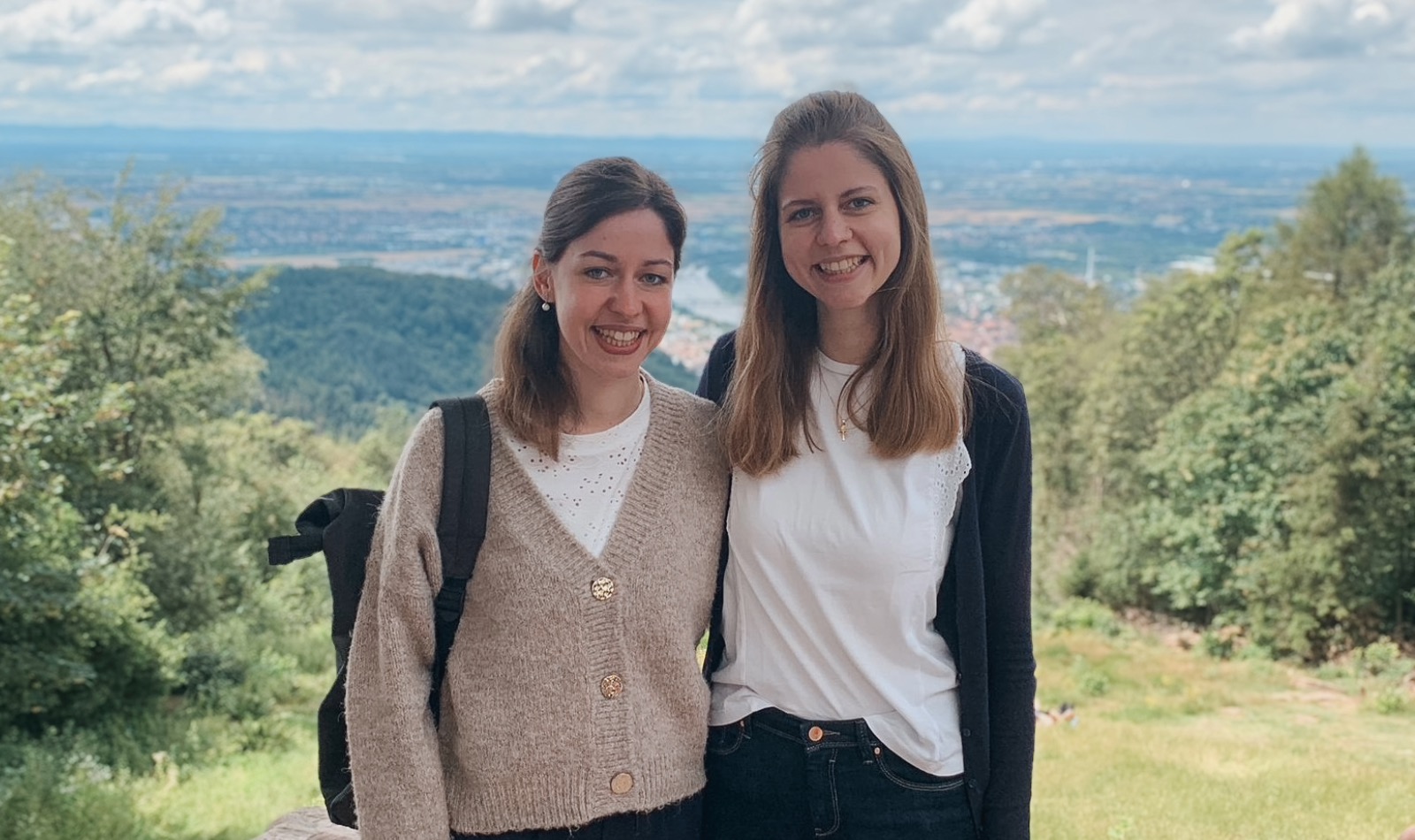Zwei junge Frauen mit langen glatten brünetten Haaren an einem Aussichtspunkt vor grüner Landschaft