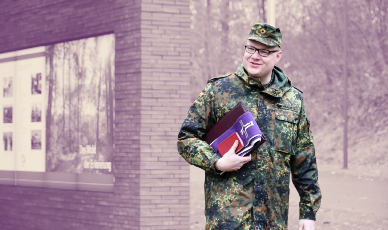 Etwas kräftigerer deutscher Bundeswehrsoldat in Uniform und Brille mit lila Buch für Seelsorge unter dem Arm