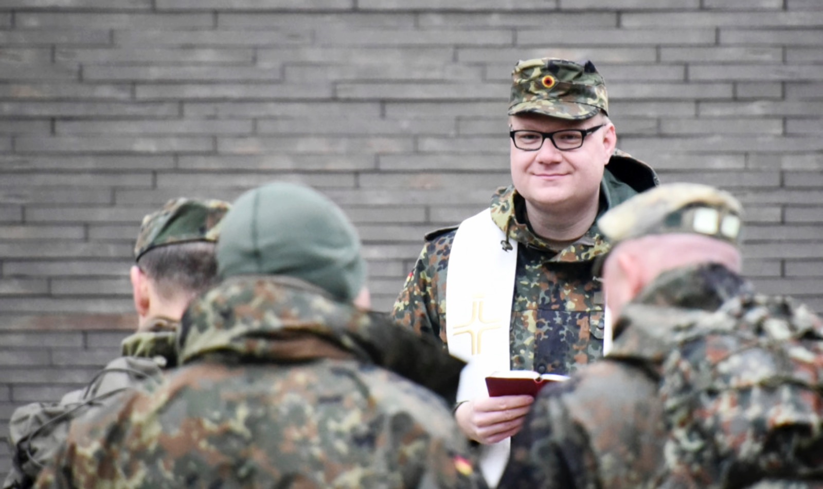 Etwas kräftigerer deutscher Bundeswehrsoldat mit Stola über der Schulter steht vor Ziegelwand und Soldaten mit Buch in der Hand