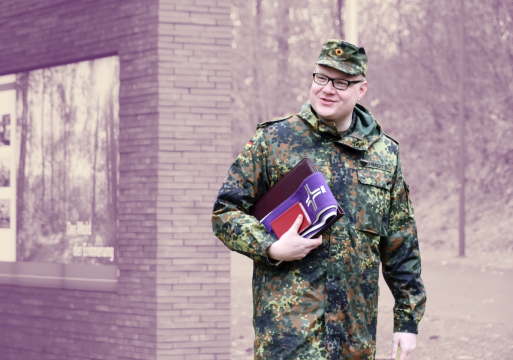 Etwas kräftigerer deutscher Bundeswehrsoldat in Uniform und Brille mit lila Buch für Seelsorge unter dem Arm