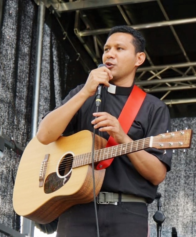 Junger indonesischer Priester in schwarzem Hemd und Priesterkragen mit Mikrofon und Gitarre auf einer schwarzen Bühne