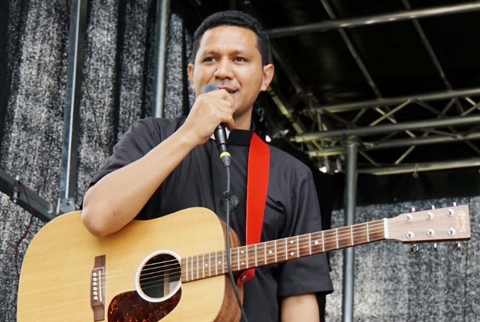 Junger indonesischer Priester in schwarzem Hemd und Priesterkragen mit Mikrofon und Gitarre auf einder schwarzen Bühne
