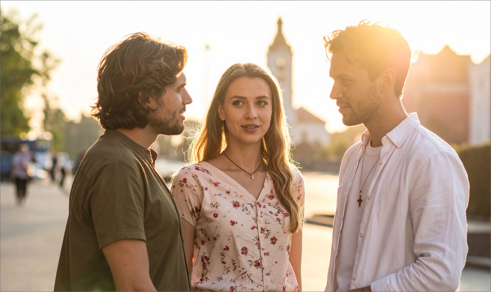 Zwei junge Männer und eine junge Frau stehend und sich unterhaltend im Freien sonmmerlicher Kleidung. Einer der Männer mit Kreuzkette. Im Hintergund eine Kirche und Sonnenstrahlen.