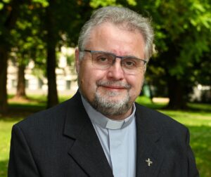 Europäischer Priester Mitte 50 mit Priesterkragen und Metallkreuz am Anzug, mit Brille, grauem Bart und grauen Haaren lächelnd vor grünem Hintergrund