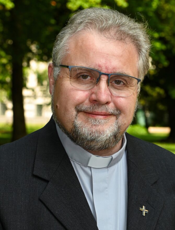 Europäischer Priester Mitte 50 mit Priesterkragen und Metallkreuz am Anzug, mit Brille, grauem Bart und grauen Haaren lächelnd vor grünem Hintergrund