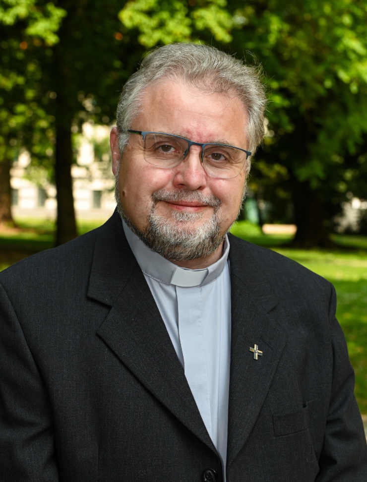 Europäischer Priester Mitte 50 mit Priesterkragen und Metallkreuz am Anzug, mit Brille, grauem Bart und grauen Haaren lächelnd vor grünem Hintergrund