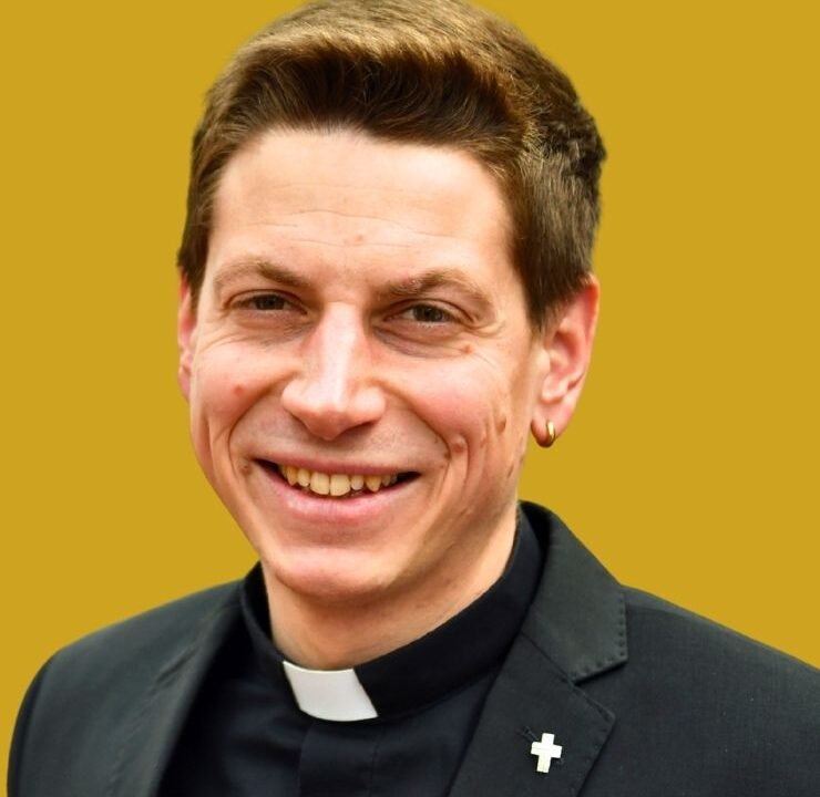 Mann lächelt, trägt schwarzes Hemd mit weißem Priesterkragen und schwarzes Sakko mit kleinem Kreuz-Pin; goldener Ohrstecker sichtbar; steht vor einfarbigem goldgelbem Hintergrund.
