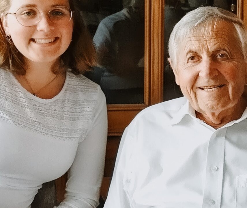 Eine junge Frau und ein älterer Mann sitzen nebeneinander und lächeln in die Kamera; hinter ihnen ein hölzerner Schrank mit Glasvitrinen, beide tragen helle Hemden.