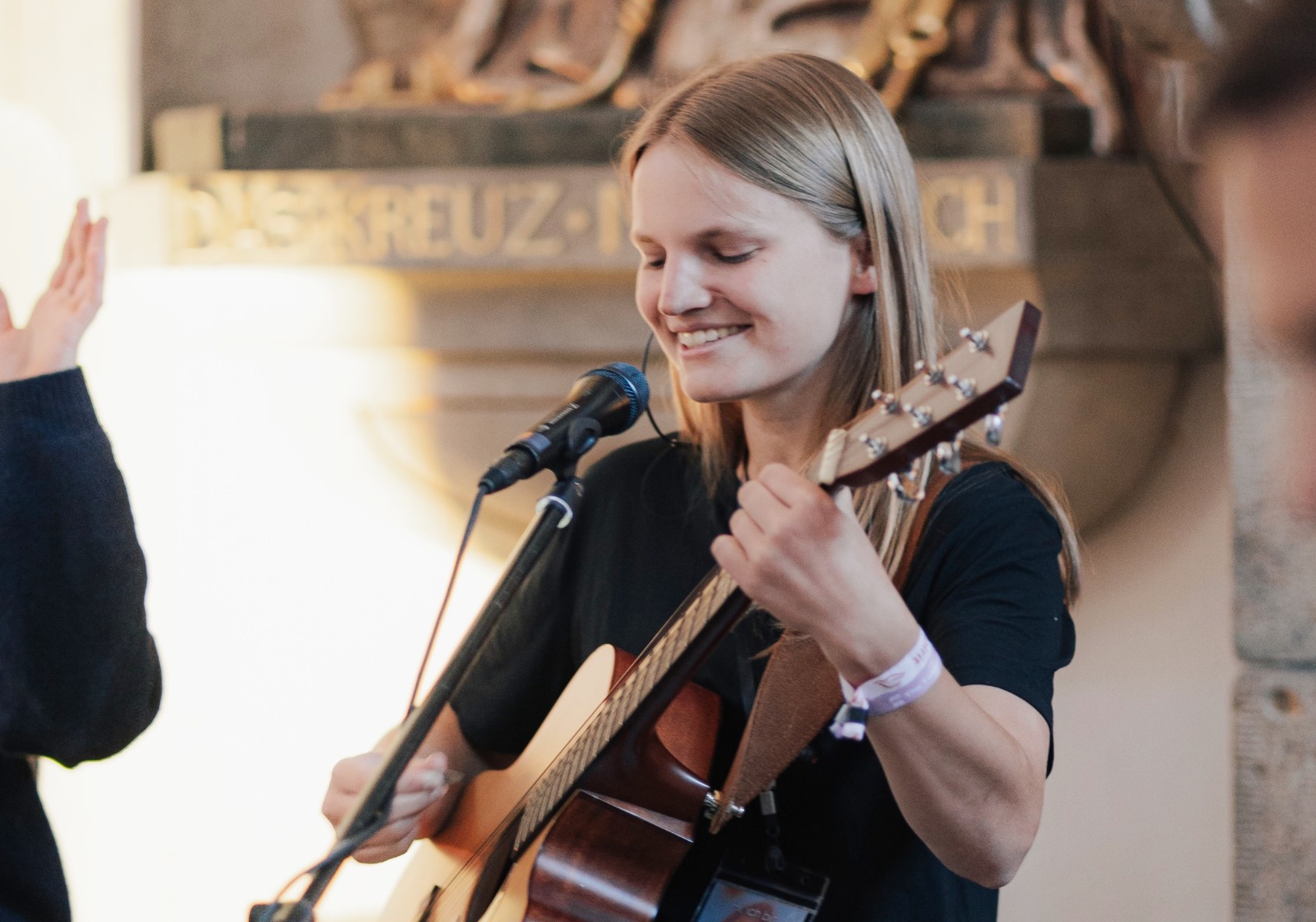 Junge Frau mit langen blonden Haaren in schwarzem T-Shirt und Armbändern mit einer Gitare beim Lobpreissingen vor einem Mikrofon in einer Kirche