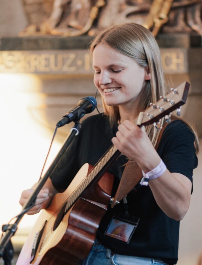 Junge Frau mit langen blonden Haaren in schwarzem T-Shirt und Armbändern mit einer Gitare beim Lobpreissingen vor einem Mikrofon in einer Kirche