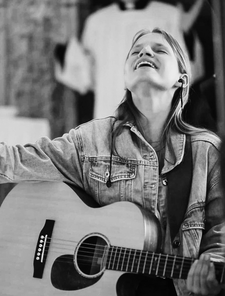 Junge Frau mit langen blonden Haaren und nach oben gerichteten Kopf in Jeansjacke mit Gitarre beim Lobpreissingen in einer Kirche