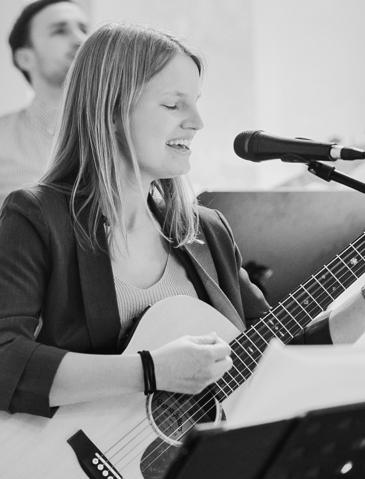 Junge Frau mit langen blonden Haaren in schwarzem Blazer mit Gitarre beim Lobpreissingen vor einem Mikrofon
