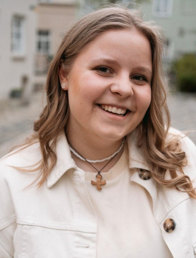 Porträt einder jungen europäischen Frau mit langen blonden gelockten Haaren, lachend mit Holzkreuzkette, weißer Kette, Ohrringen in weißem Shirt und weißem Hemd